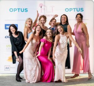 Group of women posing and celebrating at a corporate awards gala, in front of an Optus and FLT sponsor banner, captured by Light Planning Media’s event photography services.